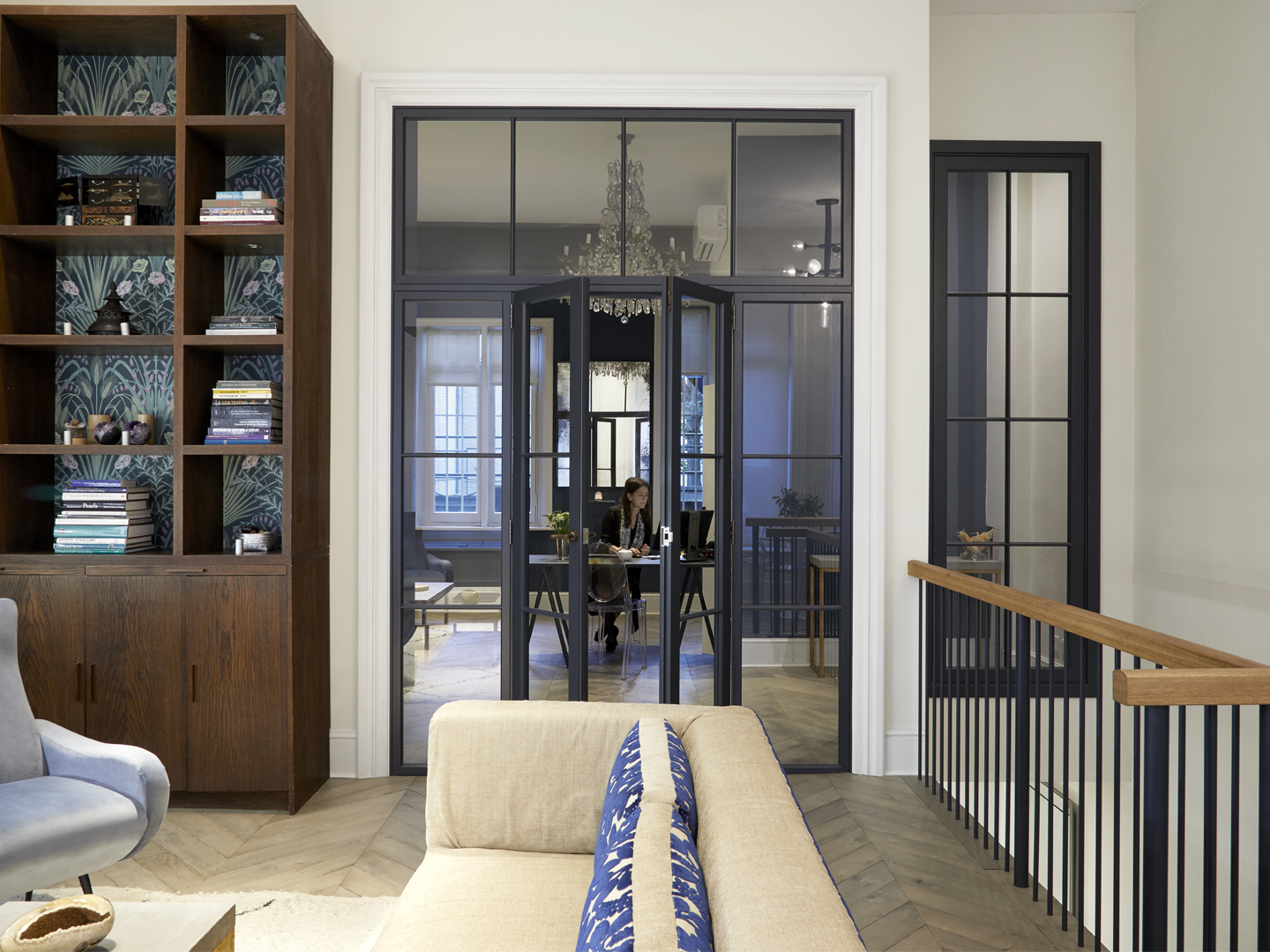 Modern living room opening through glass doors to a home office with a person working at a desk.
